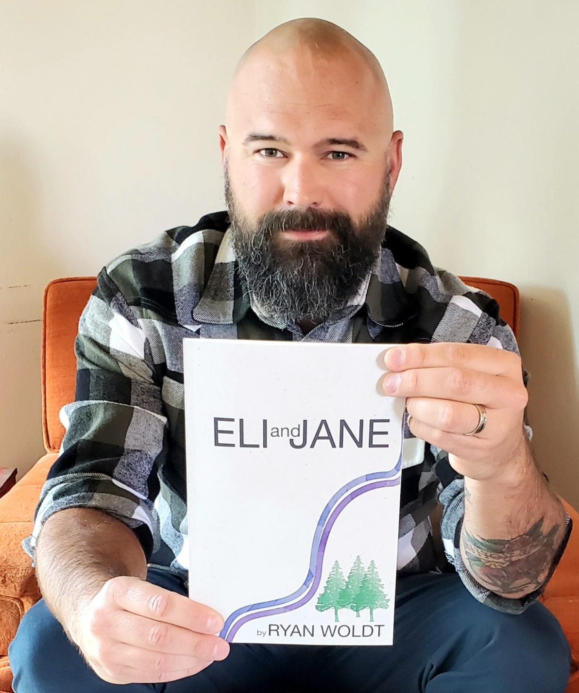 A bald, bearded, white man wearing a black and white checked flannel, seated on an orange lounge chair holding a copy of a paperback book up, titled Eli & Jane.