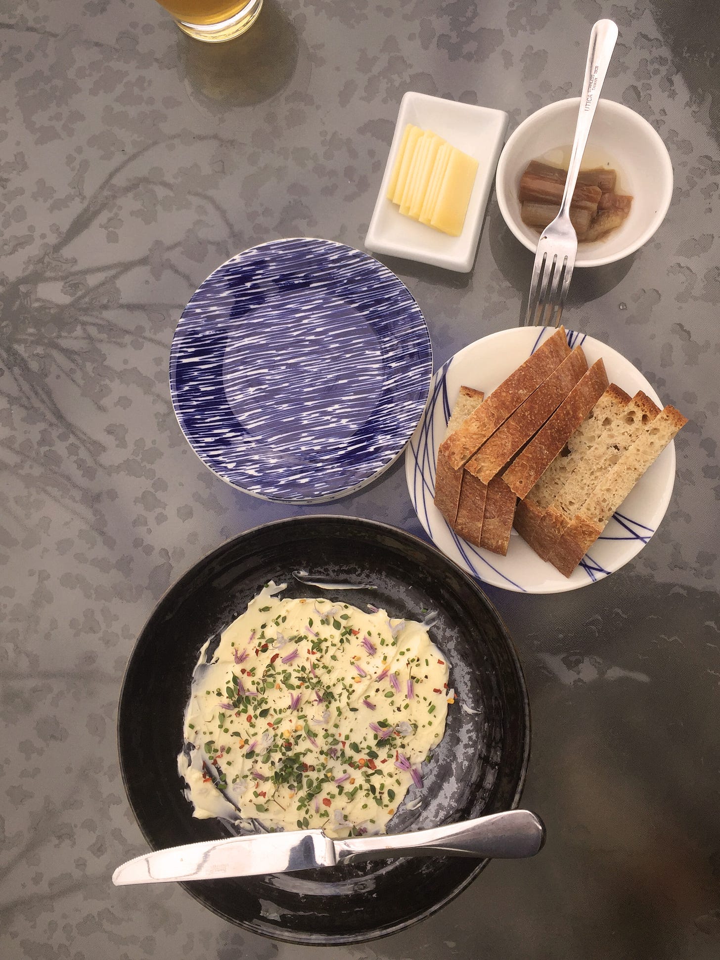 A glass patio tabletop with an arrangement of small dishes of pickles, cheese, slices of bread, and a black shallow bowl spread with butter and dusted with green herbs and small purple blossoms. 