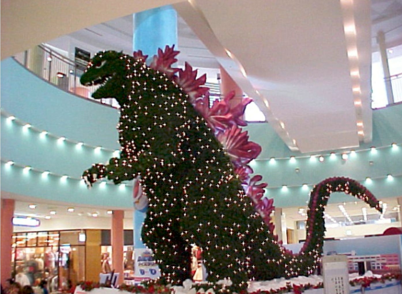 Photograph of a multi-story Christmas Tree in the shape of Godzilla adorned in fairy lights.