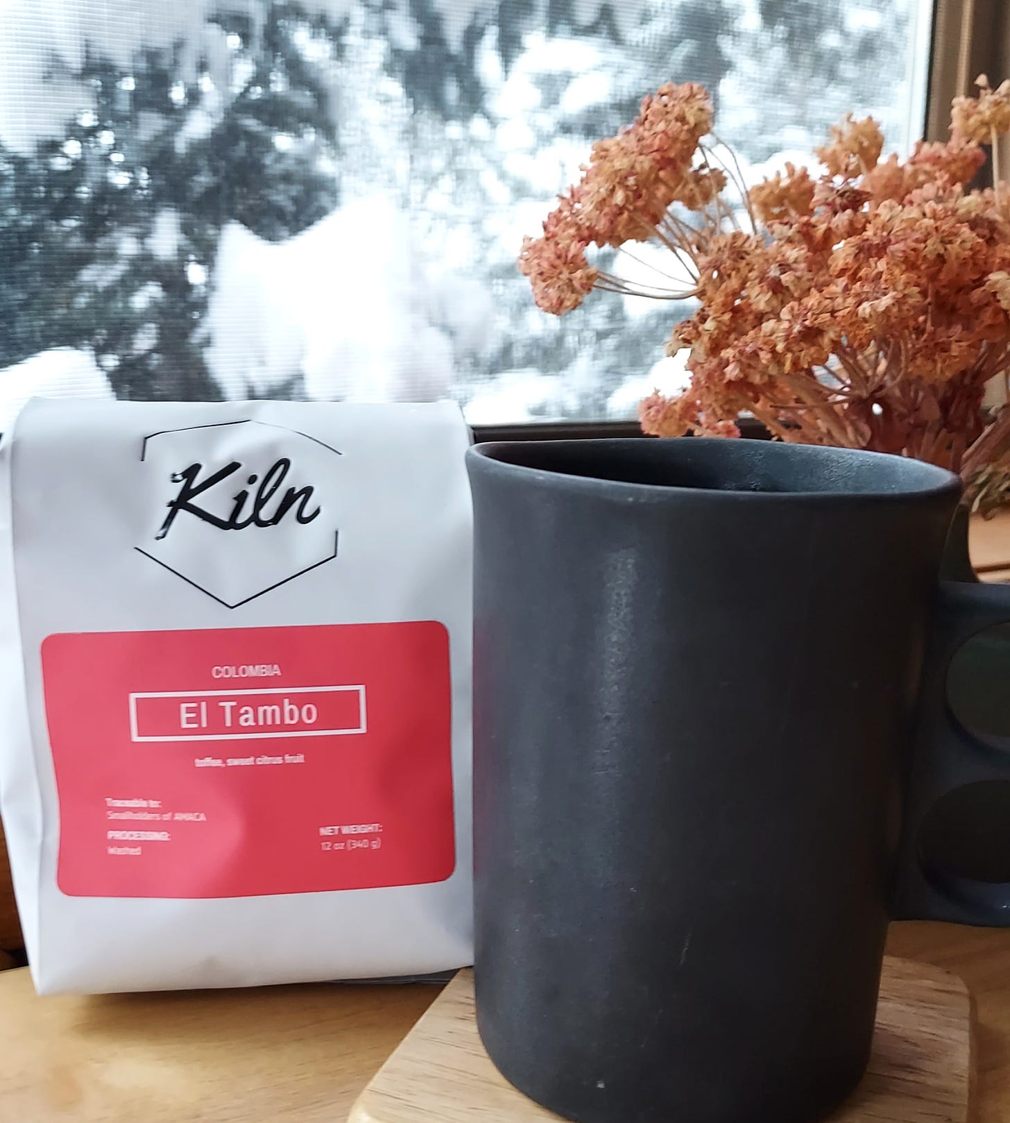 A ceramic, black coffee mug is on a wooden coaster in front of a window. Outside is a snow-covered pine tree. To the left of the mug is a white bag of coffee with a red label that says "Kiln" and in the back right is a small vase with dried, decorative flowers.