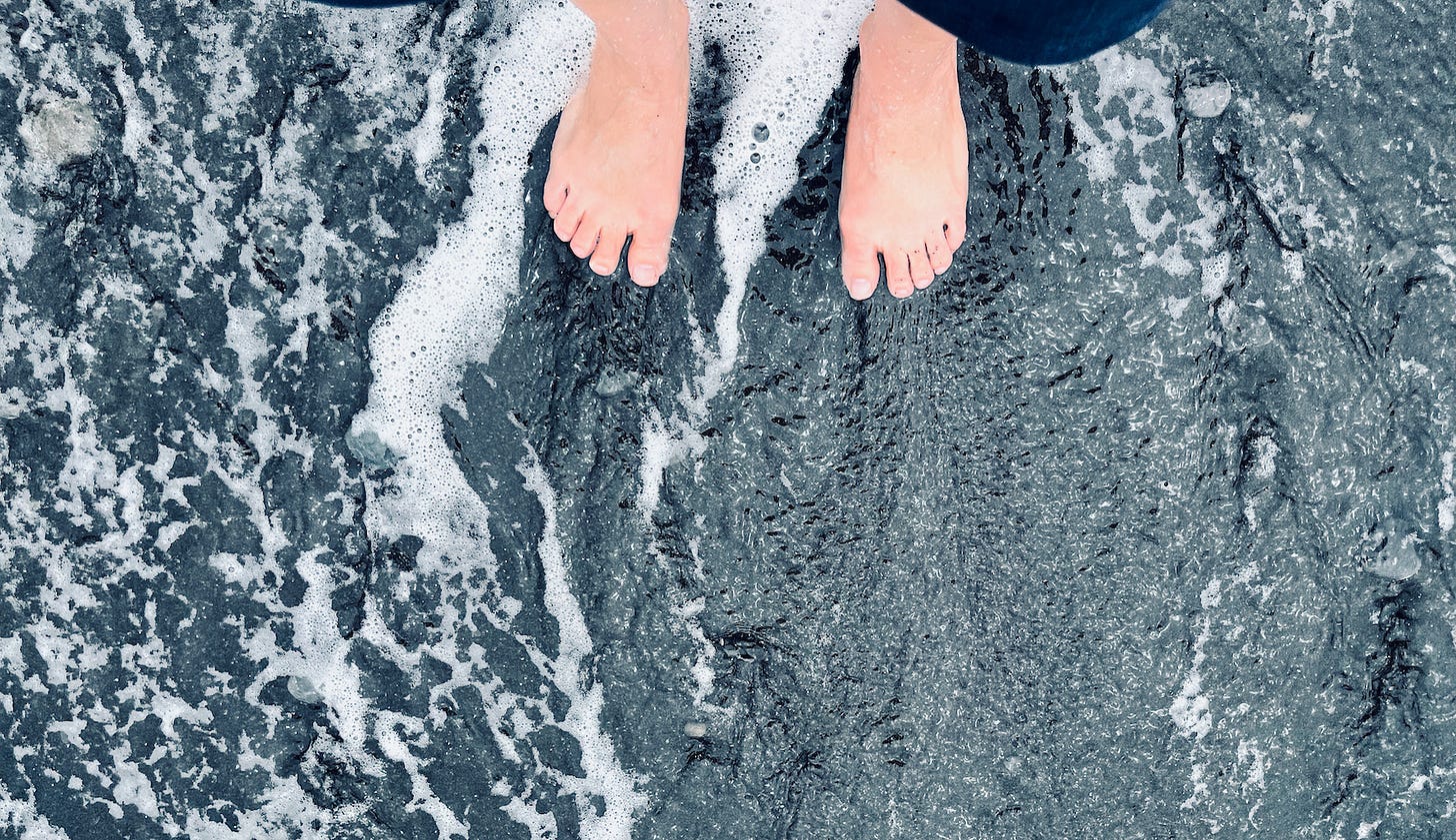A receding wave leaves lines of foam and a thin, textured sheen of water on black sand and around a light-skinned person’s feet.