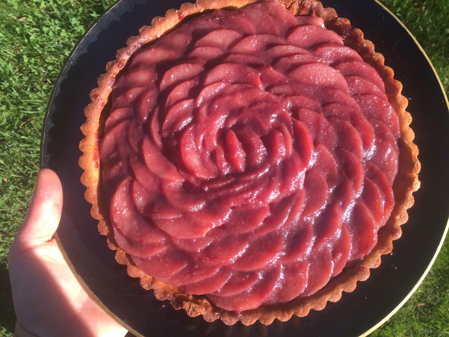 My hand holding a tart on a black platter. Deep purple pear slices are arranged in concentric, overlapping circles inside a fluted tart shell. It looks a bit like a rose.