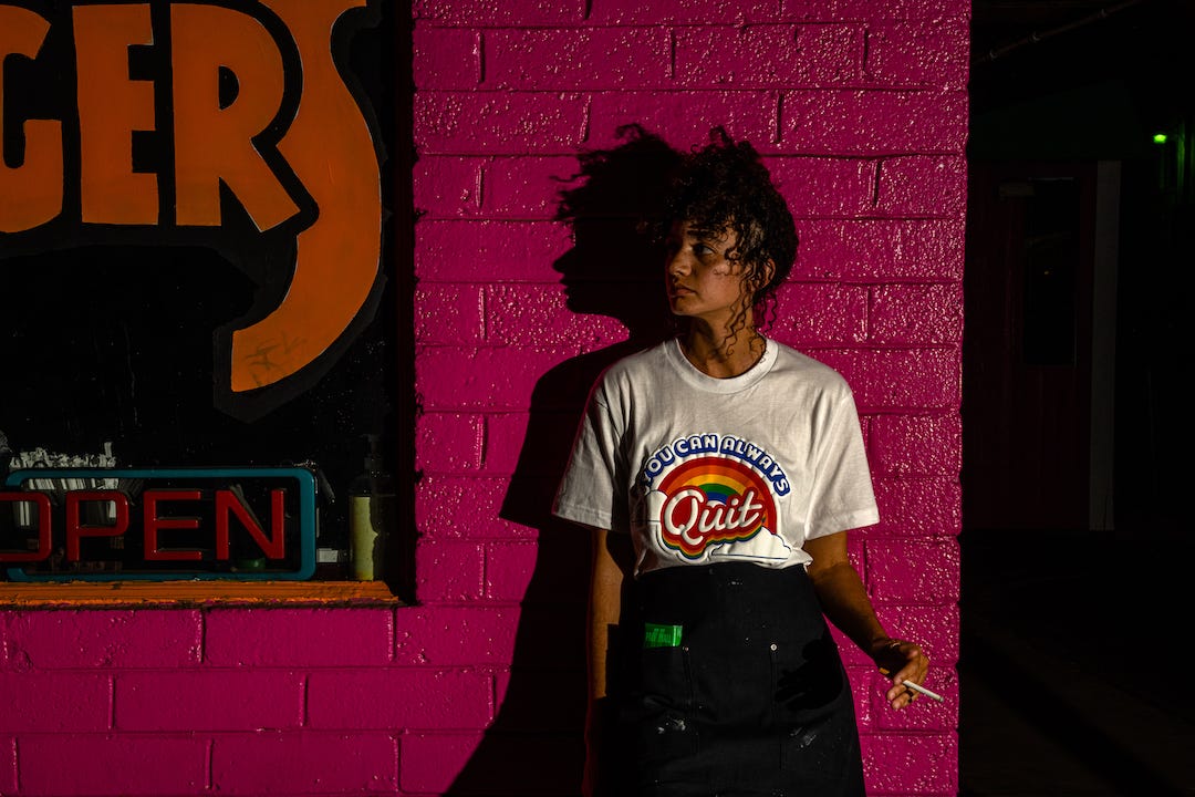 A young woman stands in front of a wet-looking purple cinderblock wall in moody lighting, wearing the You Can Always Quit t-shirt in white and a black waiter’s apron. She holds a cigarette and looks off to her right, thinking about how much she needs to quit.