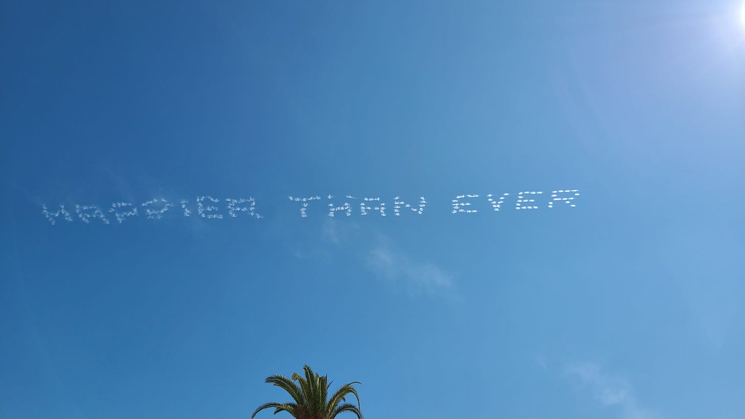 Image of skywriting seen from the VA hospital. It says, Happier Than Ever.