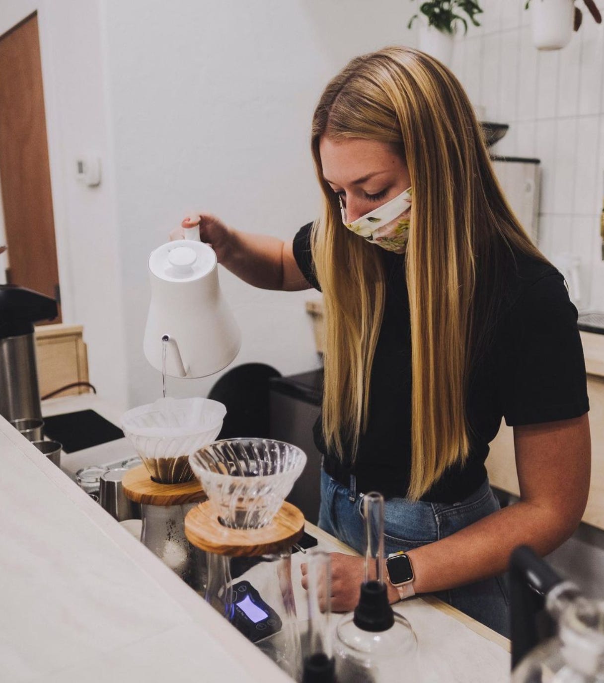 A barista pours hot water from a white goose necked pitcher into a v60 coffee brewer while standing behind a waist high bartop and in front of a white wall. She is wearing a mask over her nose and mouth and long blond hair hangs down framing her face.