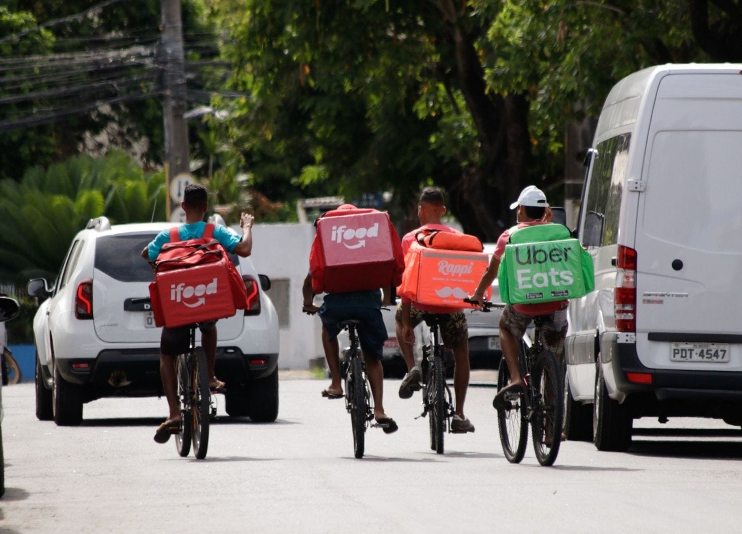 Foto de 4 entregadores no trânsito em suas bicicletas, de costas, onde se vê mochilas do iFood, Rappi e Uber Eats.