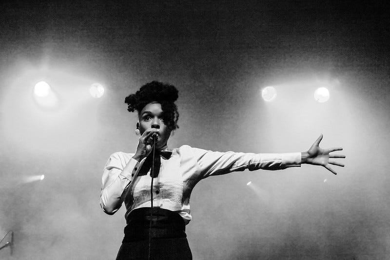 Janelle Monáe, A Black woman in a white tux shirt sings into a microphone, her right hand extended straight to the side. Stage lights glow in the fog behind her. The photo is black and white.