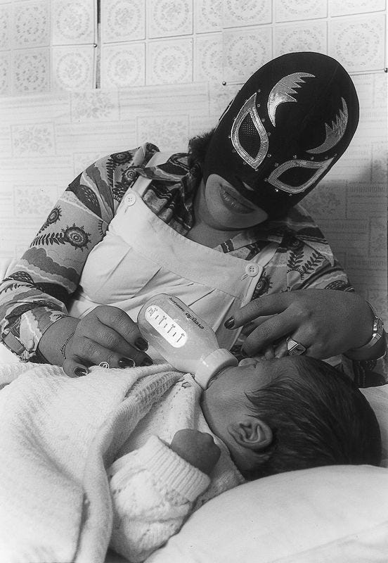 A mother wears a luchadora mask and lipstick while feeding a bottle of milk to a baby. The baby is lying down with their eyes closed.