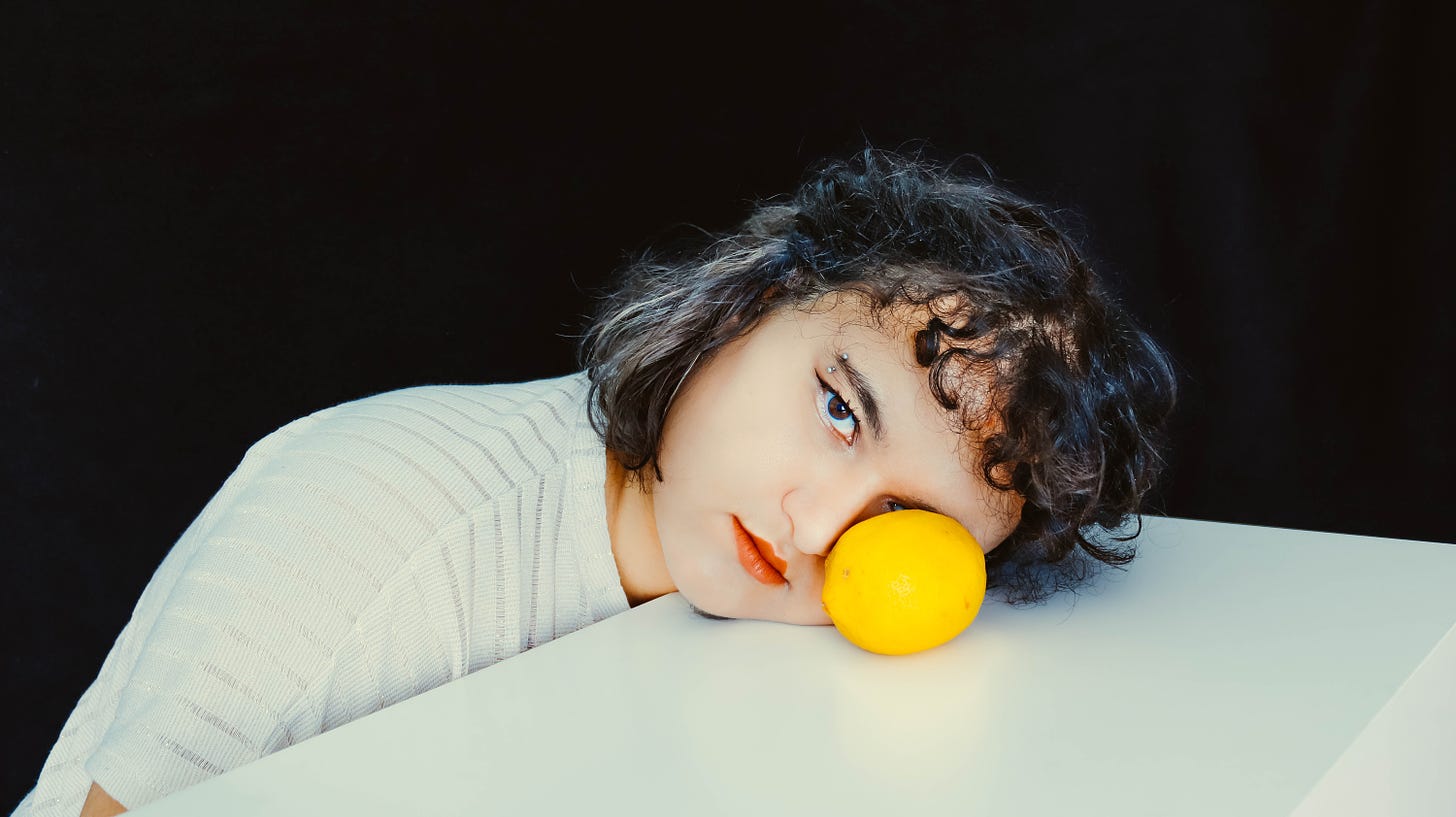 a girl with dark hair, lying her head on a table, a yellow ball is in front of one eye