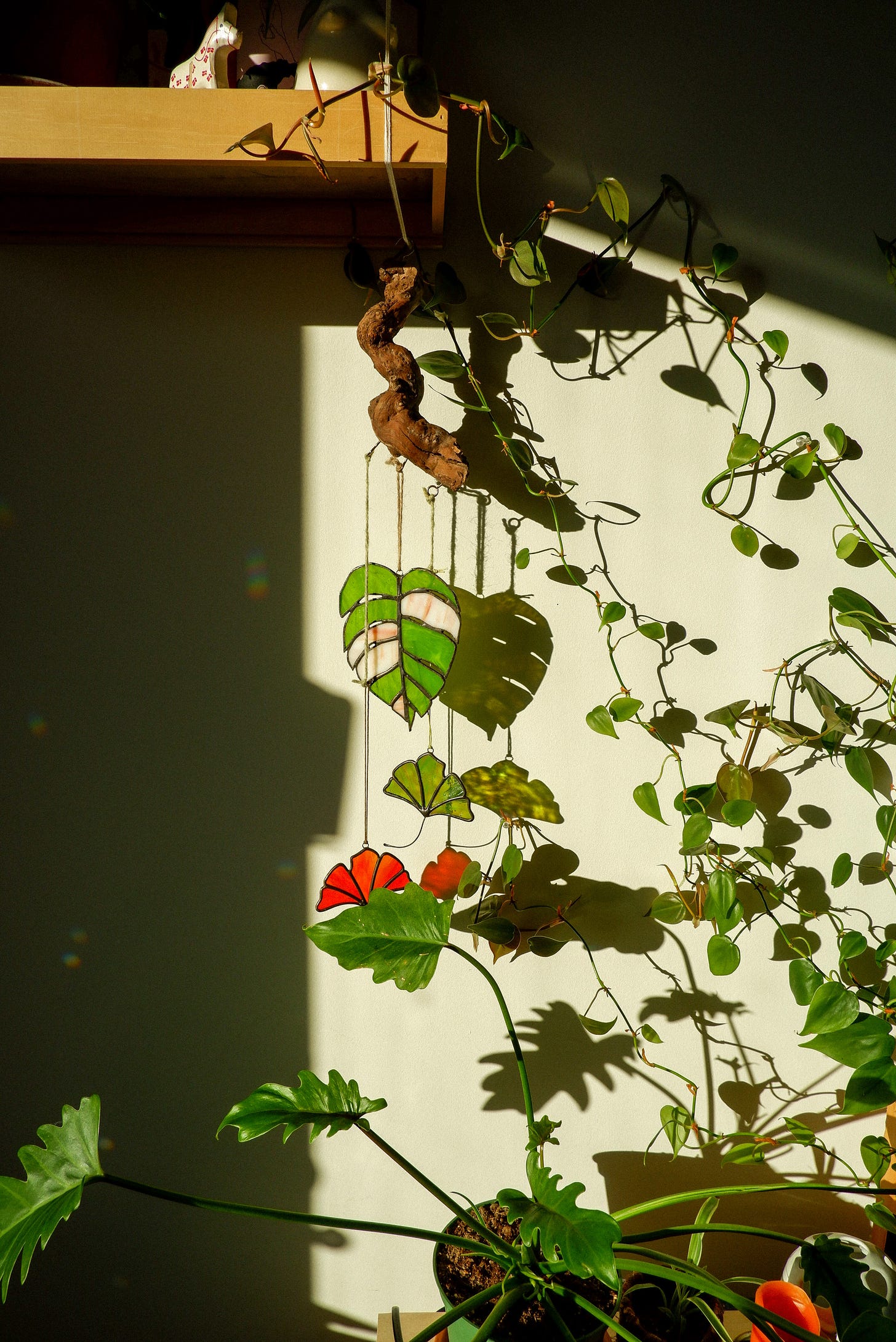 plant vines climbing a wall up to a wooden shelf. there are three stained glass leaves hanging from a stick off the shelf. the light is illuminating the vines and stained glass