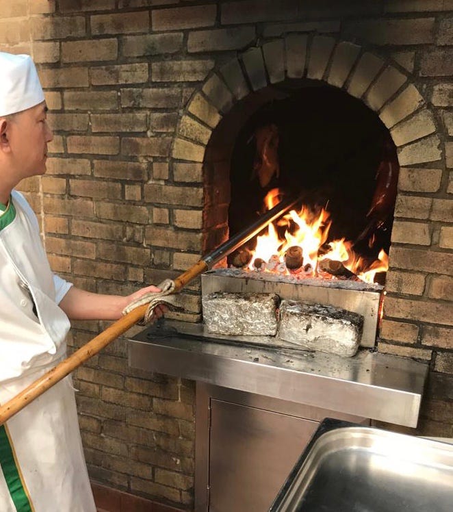 A chef stands in front of a brick oven with an open fire, wielding a long metal rod to place and retrieve ducks that are hanging inside.