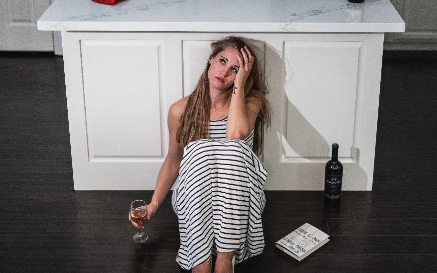 Woman in stripped dress sitting on the floor with a bottle of wine and her head in her hand.