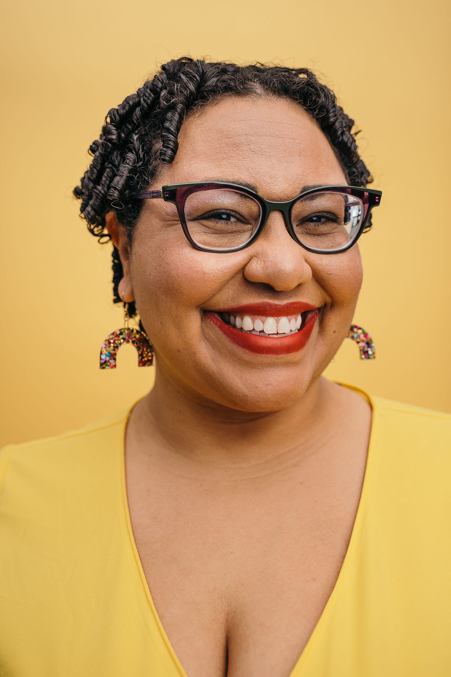 Light-skinned, short-haired Black woman smiling. She is wearing a yellow top, red lipstick, brown eyeglasses and glittery rainbow earrings. Paulius Musteikis Photography