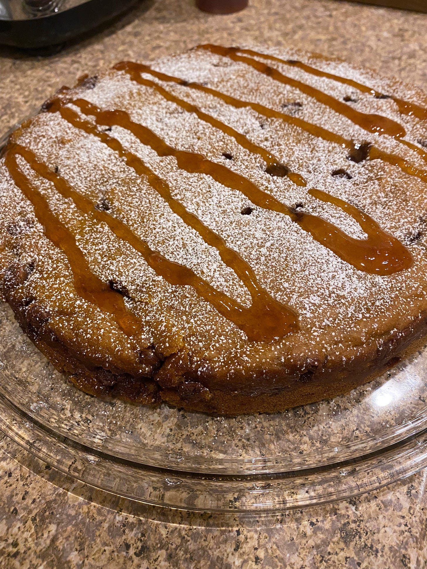 On a clear glass cake plate, a large round single-layer pumpkin cake with chocolate chips. On top is a dusting of powdered sugar and a drizzle of apple syrup.