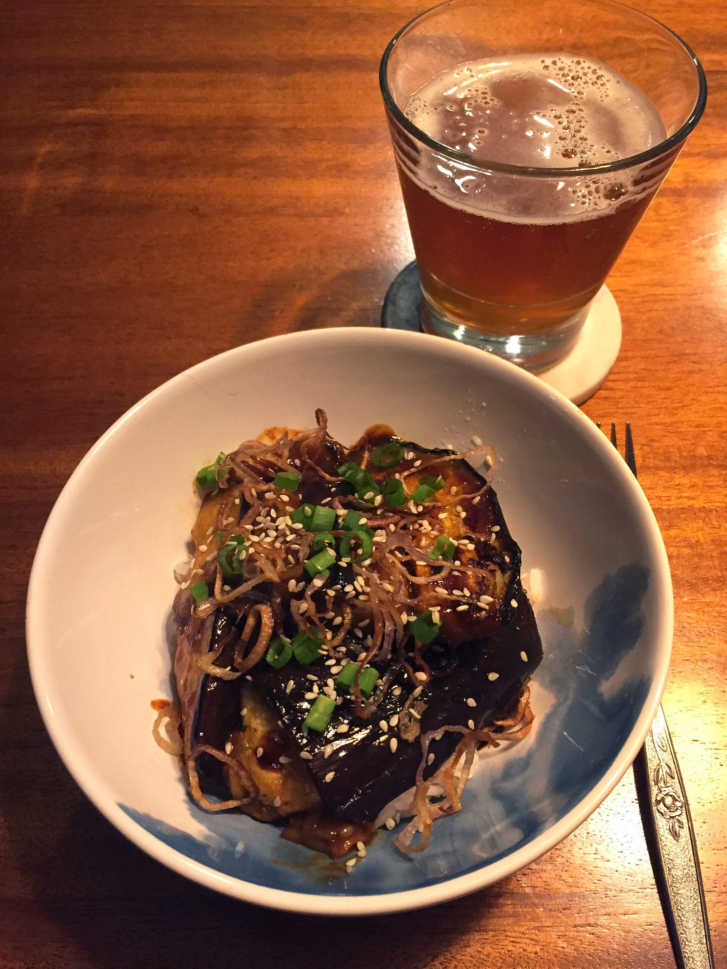 In a blue and white bowl, large slices of glazed eggplant sit on a bed of rice with fried shallots, chopped green onion, and sesame seeds scattered overtop. To the upper right of the bowl is a glass of pale ale.