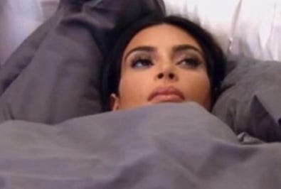 Kim Kardashian in bed, grey covers pulled up to her chin. She's staring at the ceiling looking exhausted and over everything