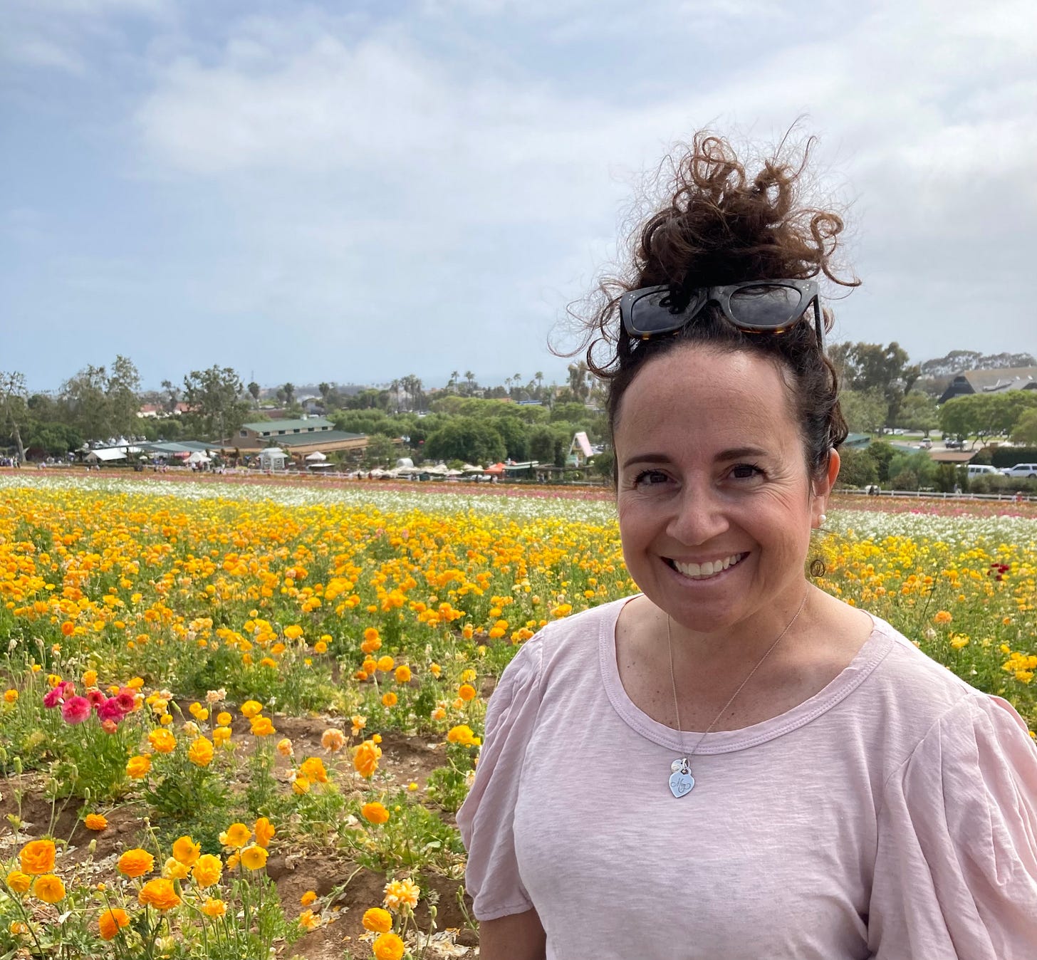 A portrait shot of a white woman with brown curly hair tied in a loose bun high on her head stands smiling to the right side of the image, with a colorful field of flowers in the background. The woman wears a pink shirt, with sunglasses propped on top of her head. She is wearing a silver pendant necklace and has brown eyes.