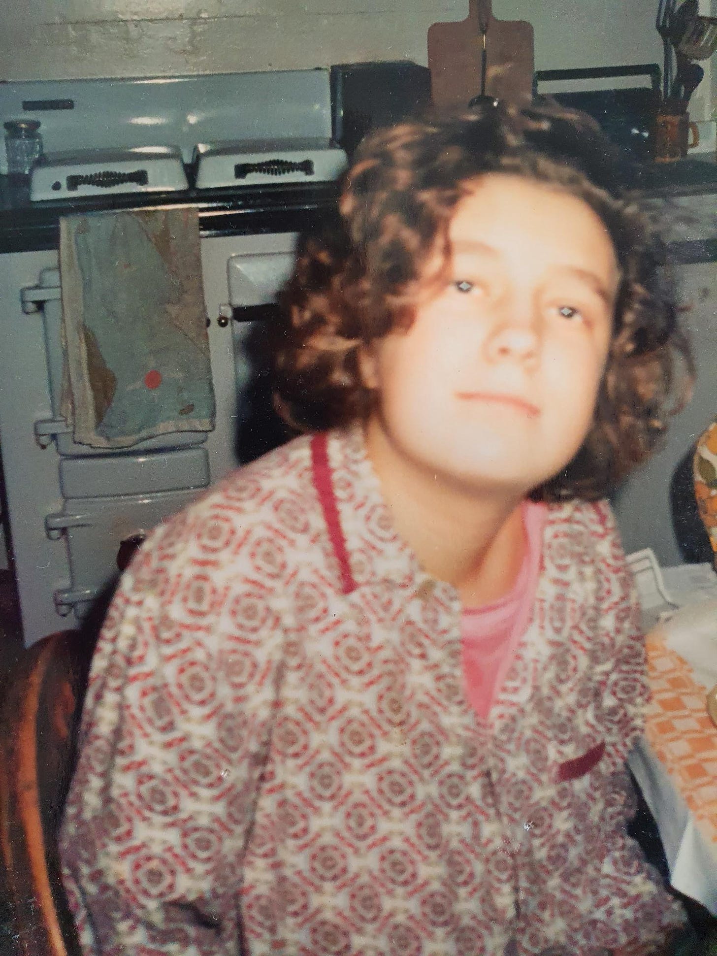 Photo of me aged 17, at the breakast table, wearing a pink t-shirt and a pair of my dad's pyjamas. I am staring into the camera with a vaguely faraway look. 
