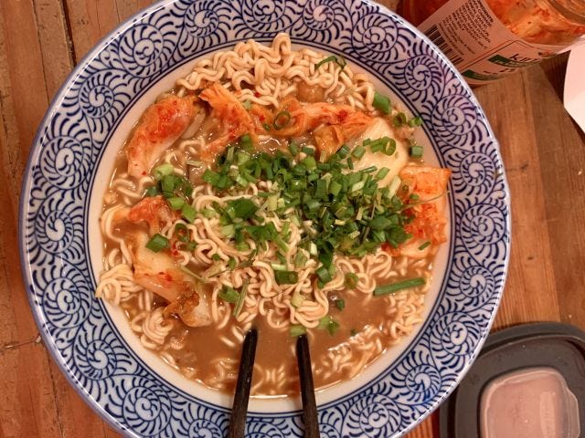 A ramen bowl with curly noodles in a soup, orange kimchi pieces and green scallions on top.
