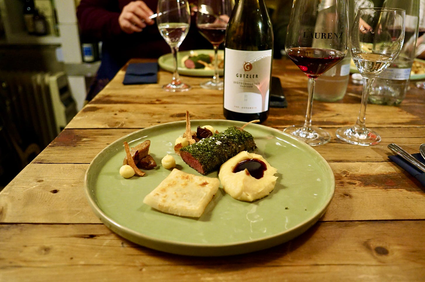 A pale green plate on a wooden table, surrounded by glasses and a bottle of wine. The food on the plate - venison and vegetables - is very neatly arranged.
