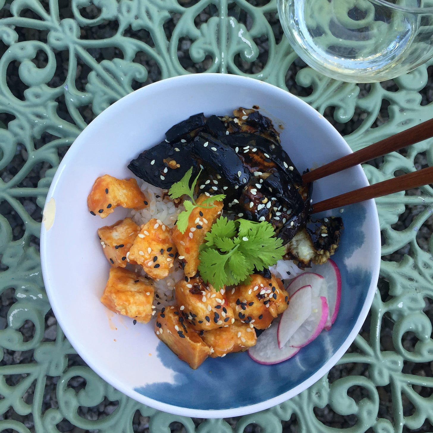 On an outdoor table, a white and blue bowl with chopsticks at one edge is filled with white rice, crispy tofu, and pieces of grilled eggplant. Everything is covered in an orangey sweet chili sauce, and radishes, sesame seeds, and cilantro garnish the top.