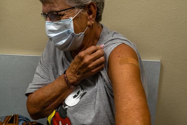 A woman in a surgical mask holds up the sleeve of her gray Mickey Mouse T-shirt to reveal a bandage where she received a booster shot.