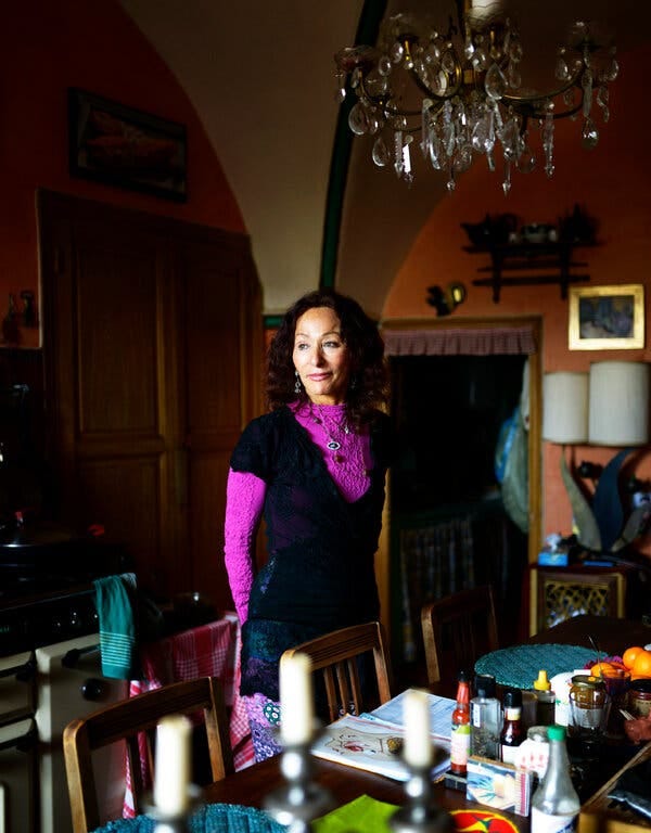 Aline Kominsky-Crumb, elegantly dressed and with shoulder-length curly brown hair, stands underneath a chandelier at a dining room table with her hands behind her back.