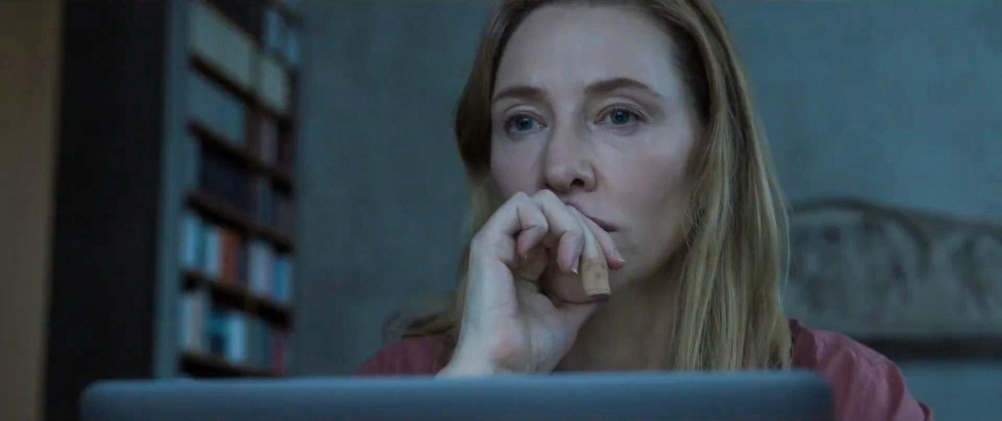 Still from Tar. Cate Blanchett sits with a laptop in front of her. She holds her hand to her mouth, looking off with an expression of anxiety. There is a band-aid on her middle finger.