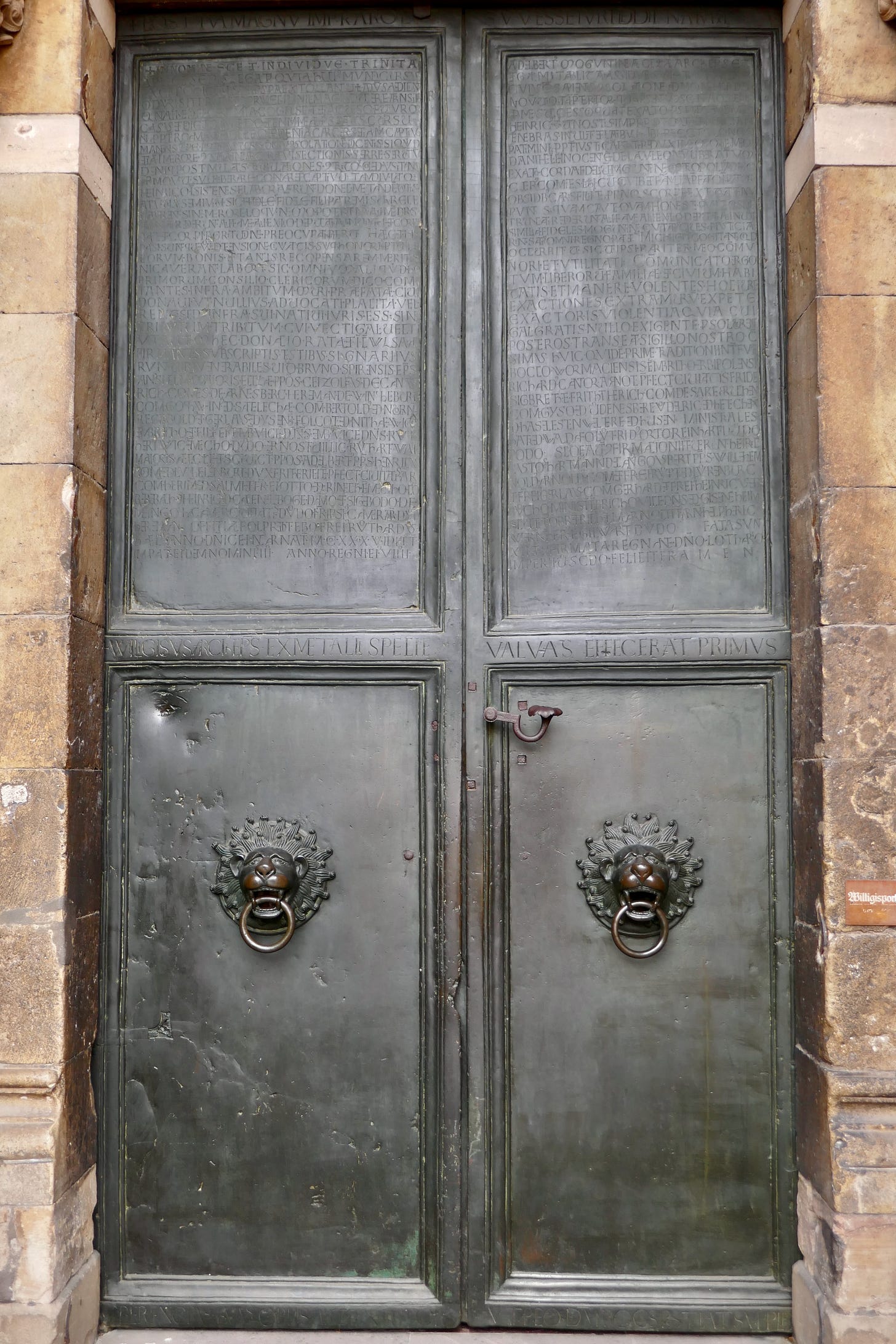 Gray steel doors with four panels and ornate door knockers on each of the bottom two panels, the brick of the structure frames the door