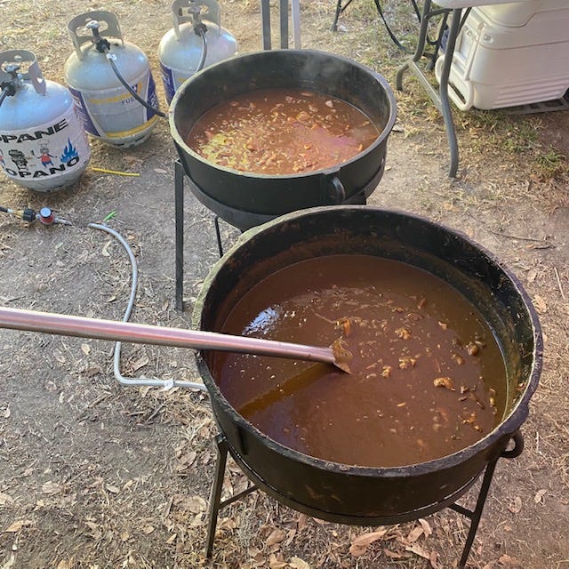 Two large outdoor cooking pots are filled with food; the rear one contains sauce piquant, a tomato-based stew, while the near one contains a dark brown gumbo. Propane tanks are visible in the background.