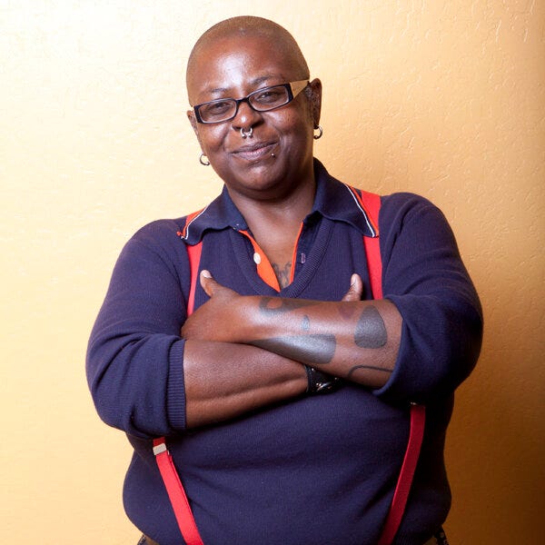 Crystal Mason looking directly into the camera and smizing. Crystal has a bald head, black rimmed rectangular glasses, and nose, lip, and ear piercings. Crystal is wearing a blue long-sleeved polo with orange trim on the collar and orange suspenders. Crystal's arms are crossed. Crystal is against a golden background. 