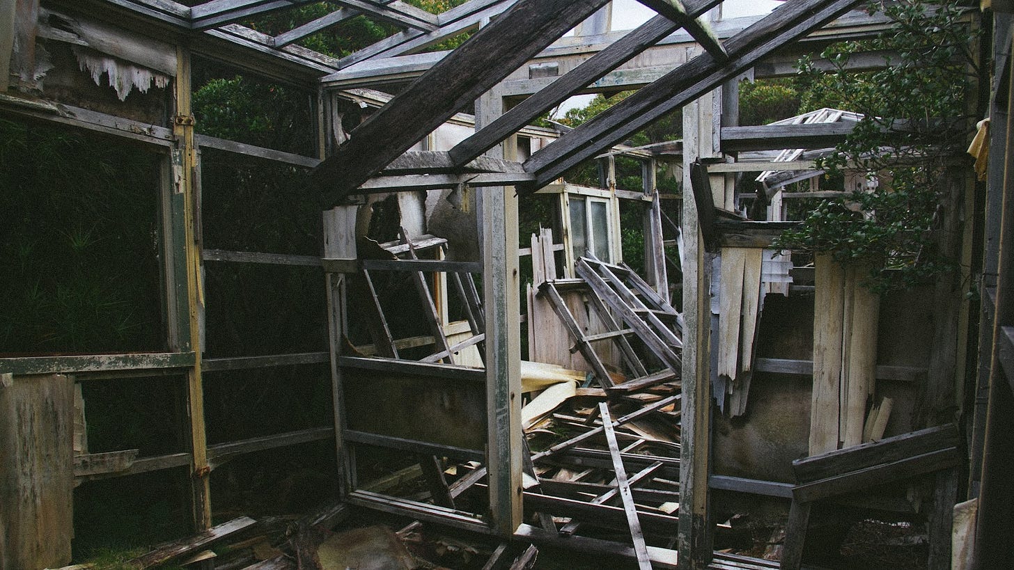 A photograph of the frame of a hut, which is now just a skeleton - with debris from the building blocking the floor and doorways. There is a tree growing through one side.