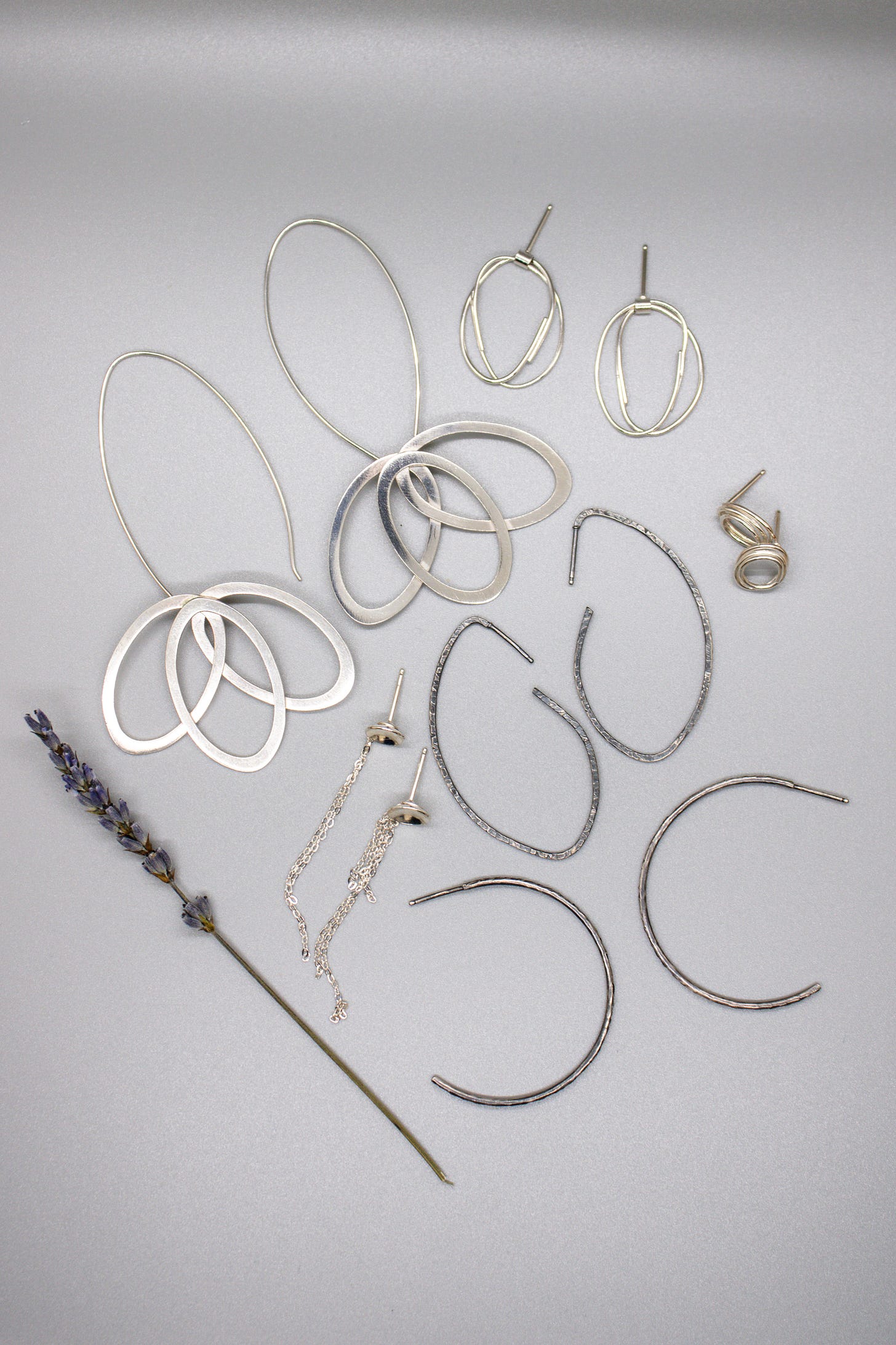 Image Description: A photo of A. Clarke Metalsmith jewelry pieces. On the bottom left is a dried stalk of lavender. From the top left to right are Stella Drops (3 ovals connected at the top, made of sterling silver pair brushed with steel wool), India's Studs (a silver circle of metal whose ends wind around the circle like arms in a hug), silver studs (tightly wound coils), Lynnelle's Hoops (large angled hoops with textured notched metal), Hope's Hoops (medium-sized traditional hoops with textured notched metal), and Lisa's Studs (silver curved disks with thin strands of chain hanging down)