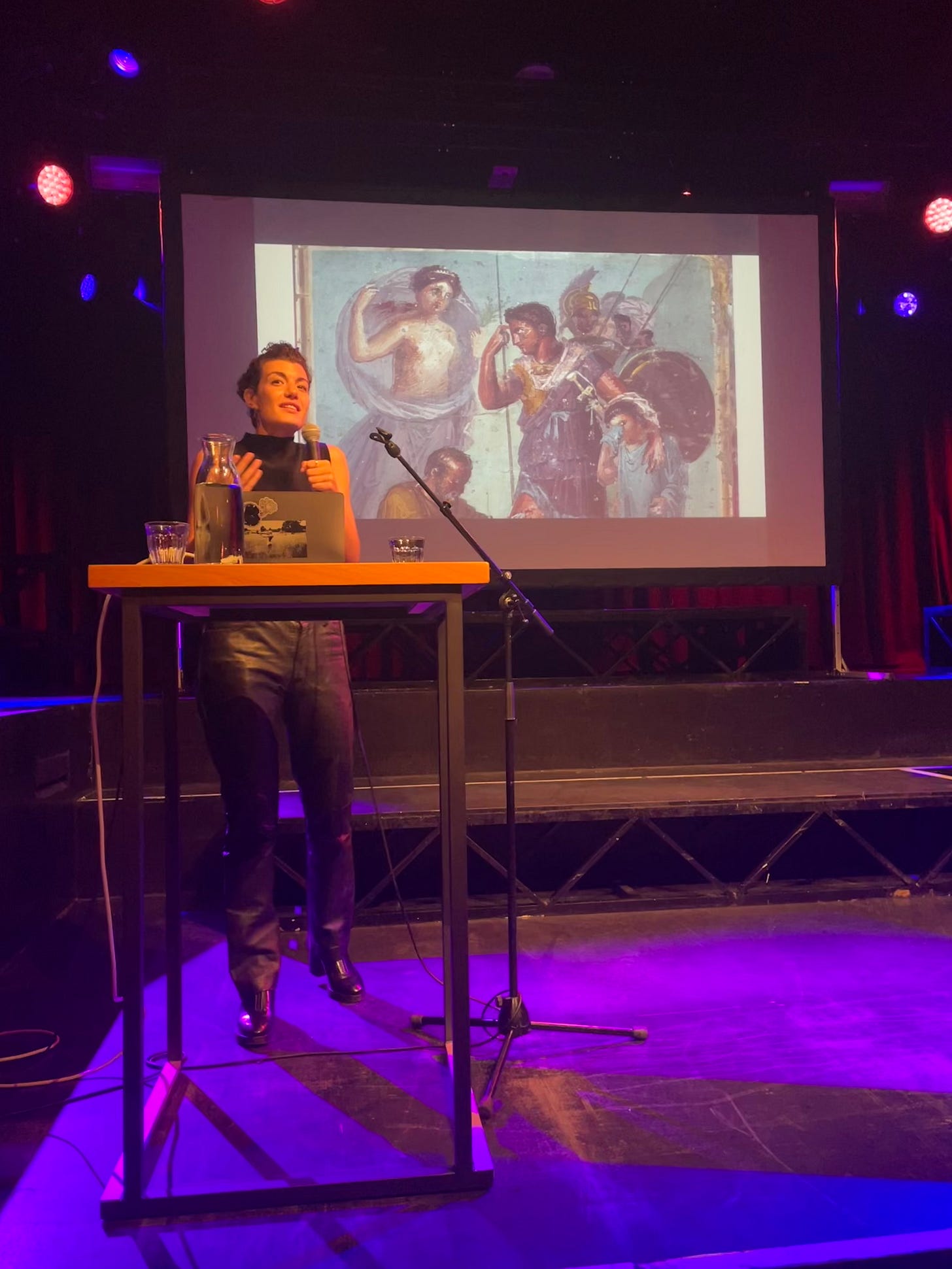 foto van Avery Trufelman op het podium achter een tafel met een laptop er op en een microfoonin haar hand. Op de achtergrond een abeelding van Aeneas