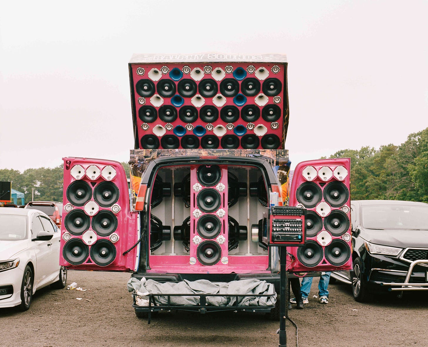 Image: The back doors of a van are open, revealing an elaborate custom bubble gum pink sound system. There are even more speakers mounted on top of the van.