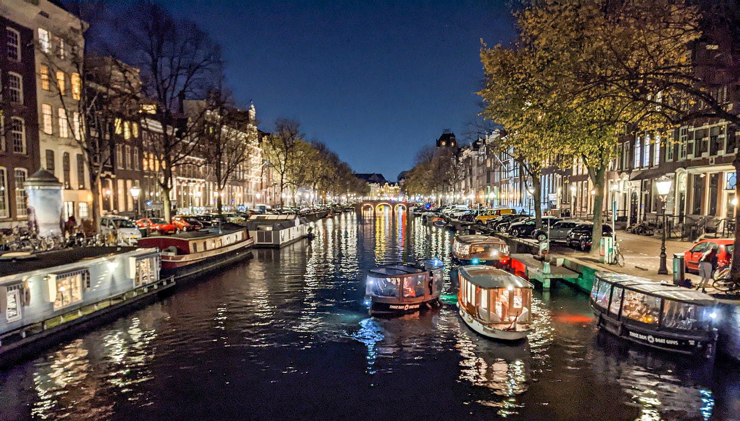 An Amsterdam canal at night with houseboats on the left, several tour boats cruising, all of it illuminated by bright white lights. 