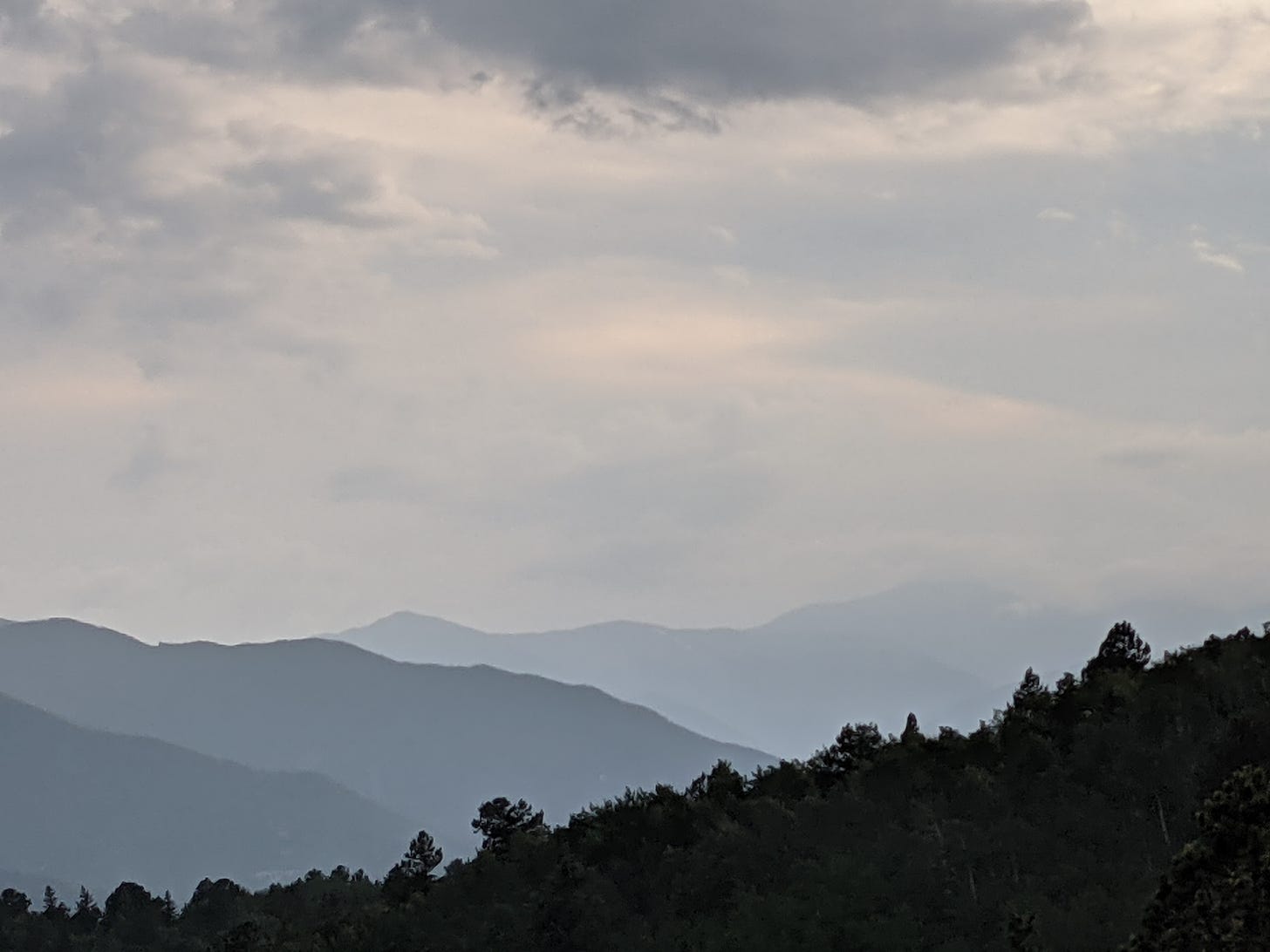 A view of blue layered mountains with low blue and white clouds. There is a steep hill of dark green trees in the foreground. 
