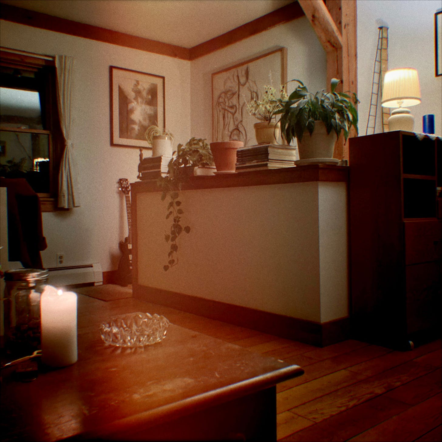 A snapshot of a lounge area with a sepia hue. In the lower right corner is a dark brown wooden table with a lit white candle and a cut glass bowl on it. Further to the right are windows with open curtains. In the centre, there is a high white ledge with several plants on it, behind there are two paintings on the white walls. To the top left is a lit small white lamp on another dark wooden piece of furniture.