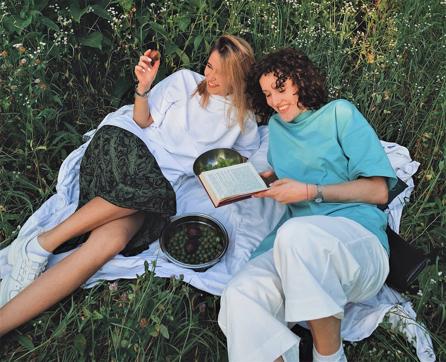 two girls laying on blanket in a green field reading a book and eating fruit