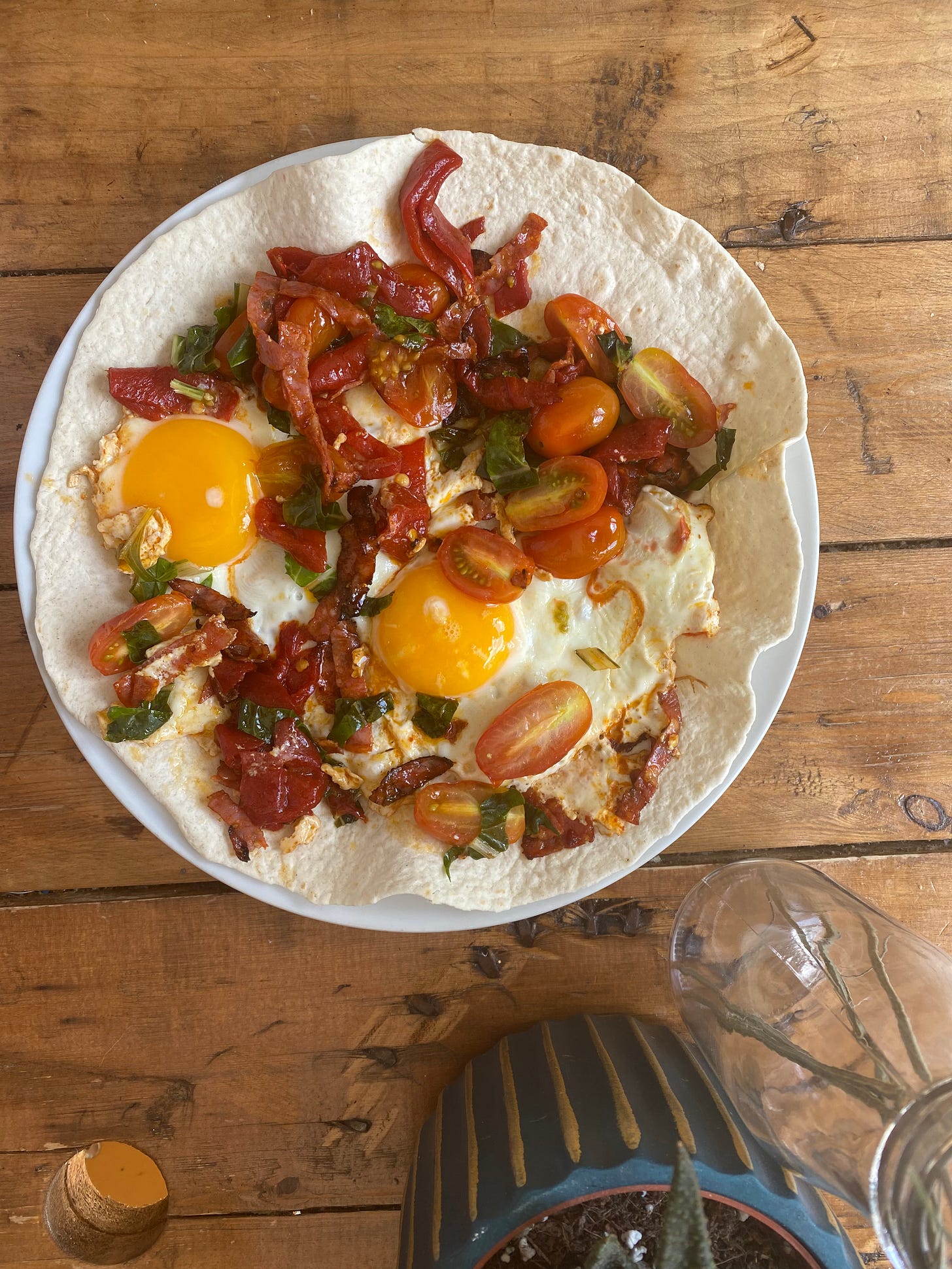 Tortilla wrap topped with two fried eggs, tomatoes, peppers and green leafy vegetable.