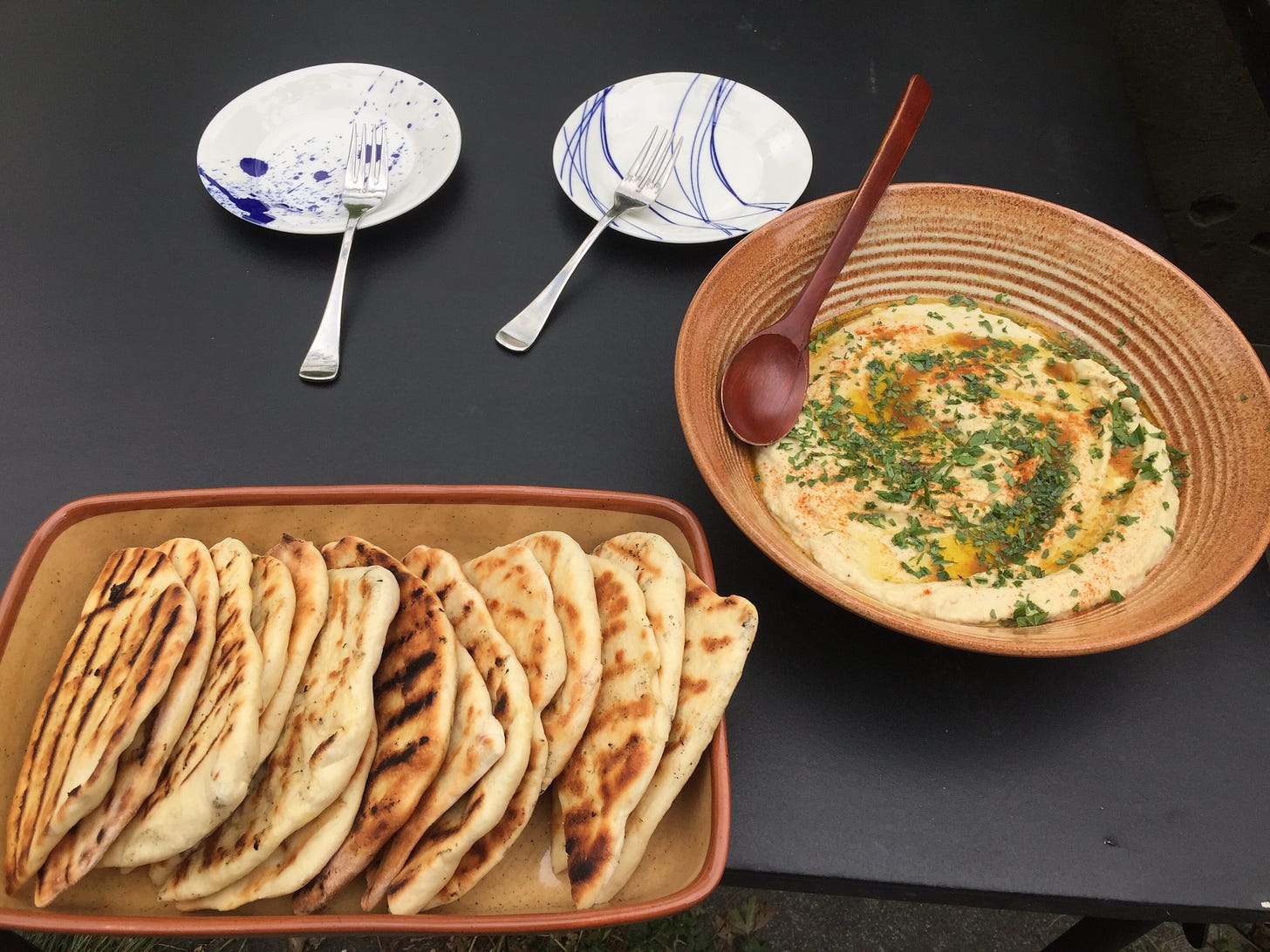 on a black table, a dish of baba ganoush sprinkled with parsley, mint, and smoked paprika, with a swirl of olive oil. Next to it is a rectangular dish full of half-moon shaped slices of grilled pita. Behind them are two small white plates with forks resting on them.