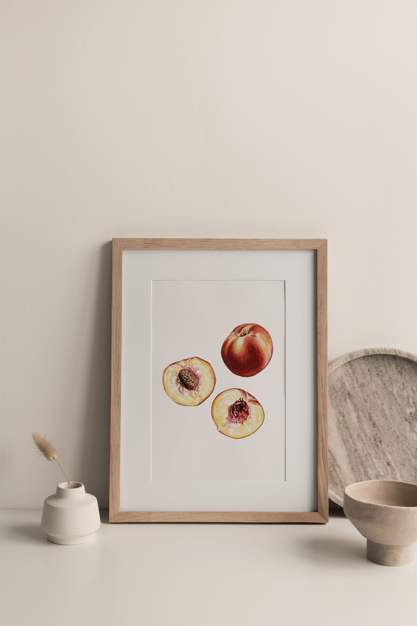 A framed peach painting, sitting on a surface with a vase to the left and a bowl to the right of it. 