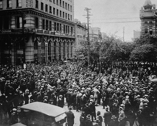A black and white photo in which a large crowd gathers in front of a bank. A car can be seen in the foreground.