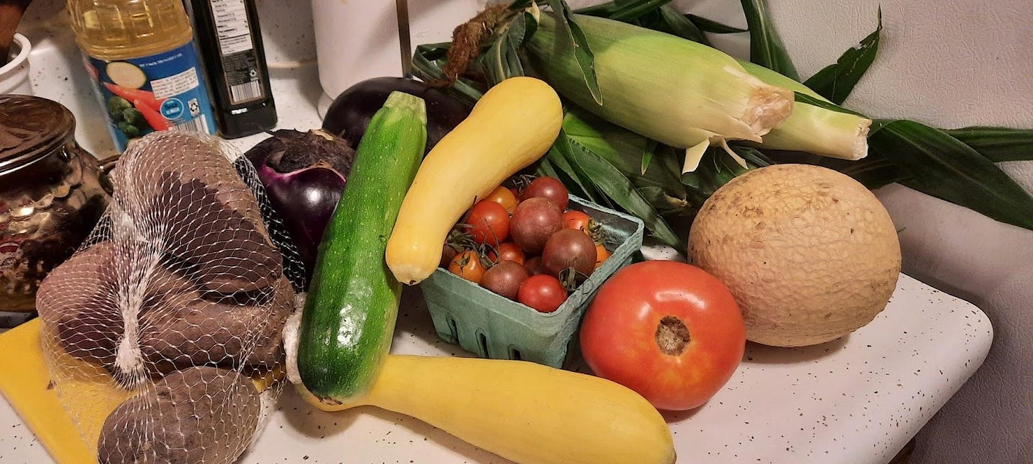 Fresh fruit and vegetables on a kitchen counter. There are potatoes, corn, eggplant, tomatoes, zucchini and a small melon.