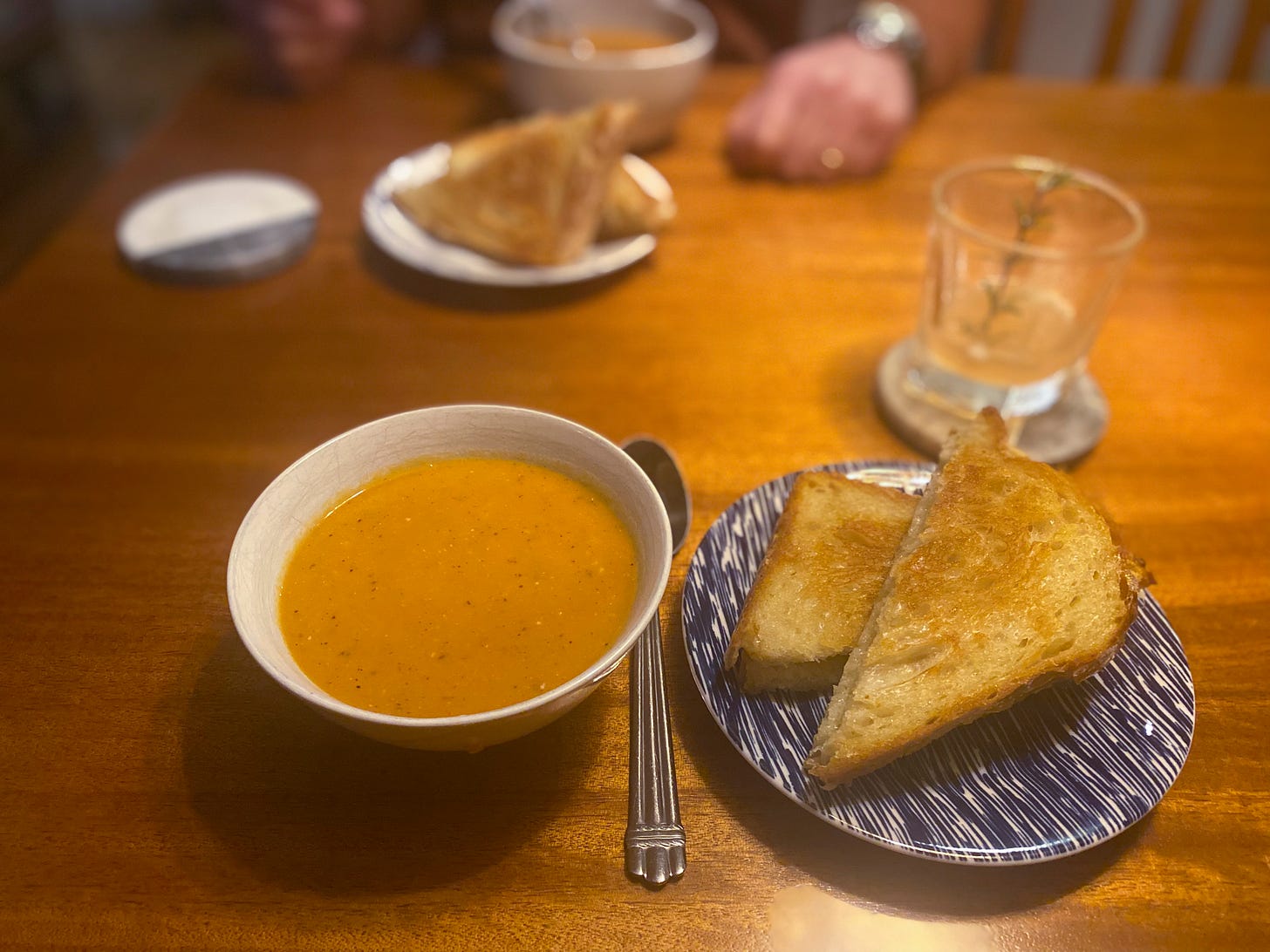 A small bowl of curried tomato and lentil soup, and a grilled cheese sandwich on a blue and white plate next to it. Across the table is the same, and a half-finished cocktail with a sprig of rosemary rests on a coaster between them and to the right.