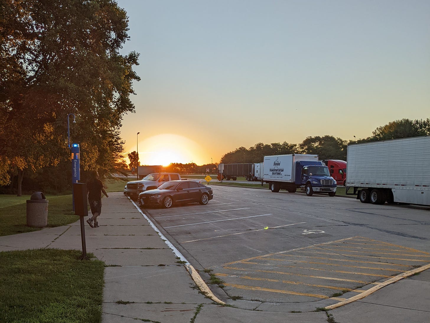 Photograph of the sun rising over a rest stop parking lot. There are several cars and semitrucks parked. The perspective is from someone standing on a sidewalk, which is partly visible. There are very large trees and and some green grass behind the sidewalk. The run is yellow-orange and the sky above it is very very light blue.