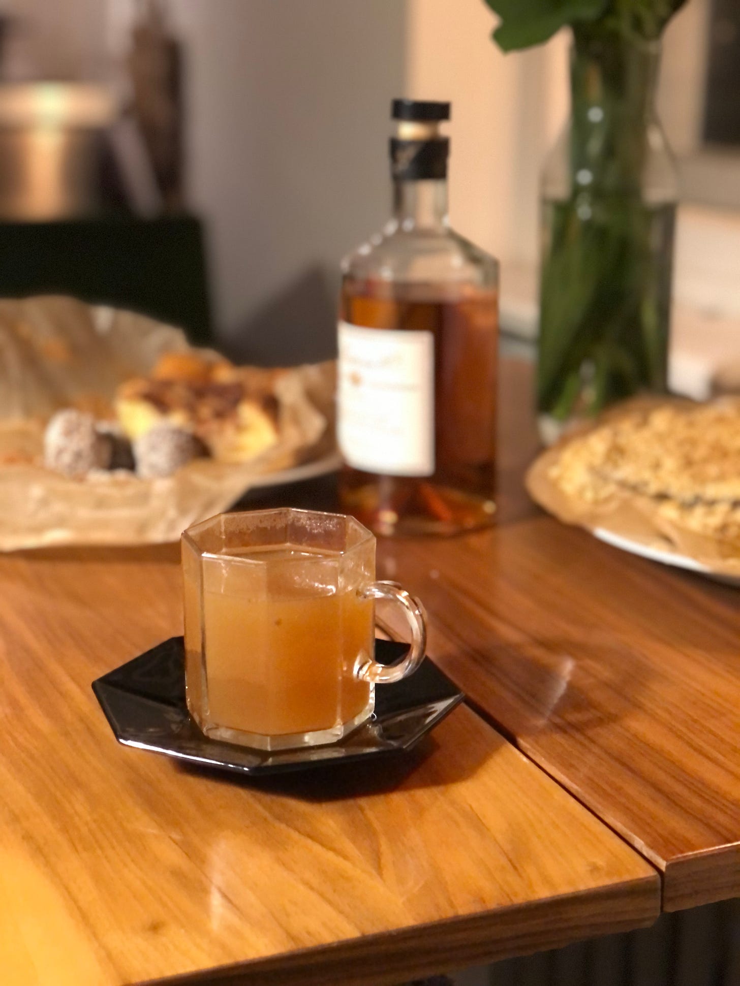 A clear octagonal short glass with a handle, on a black octagonal saucer, on the edge of a wooden table. In the glass is a cloudy amber liquid. Out of focus behind, a bottle of amber spirit, and two dishes of food. 