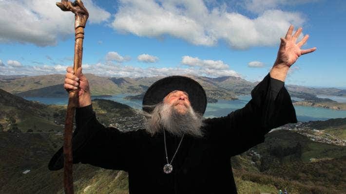 A picture of an extremely wizardly looking gent; black robe and hat, long gray beard, wizard’s staff etc, holding up his hand spellcaster-style in front of a dramatic New Zealand landscape. 