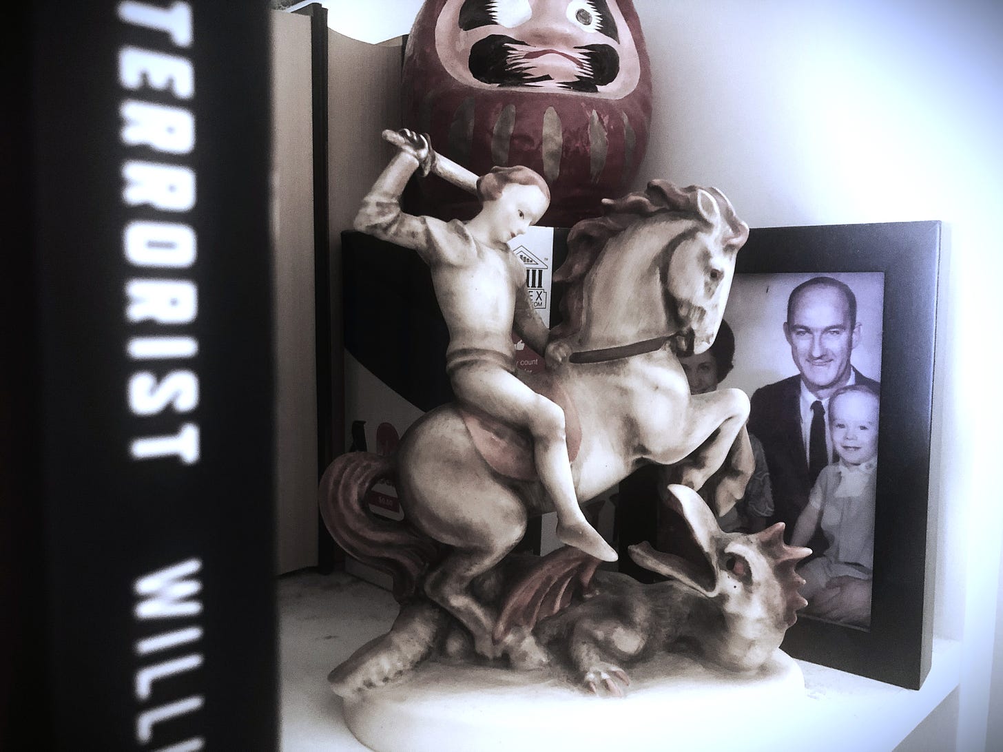 A Hummel figure of St. George on horseback fighting a dragon sits on a shelf, partially obscuring an old family photograph showing a father and son. A Japanese daruma doll sits above and behind with one eye filled in. In the foreground is the blurry spine of a book, with only the words "Terrorist Will" visible.