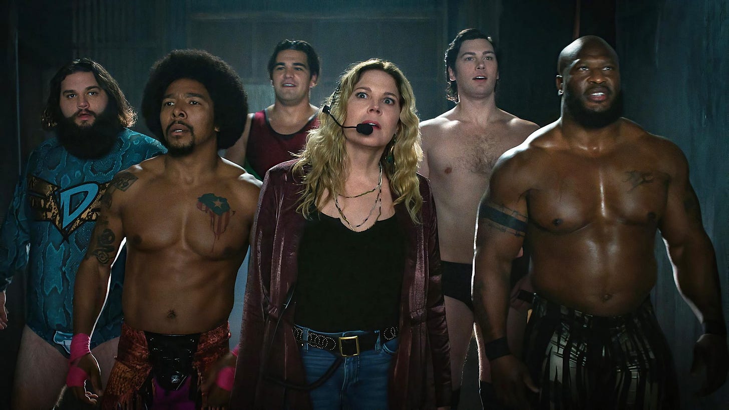 Photo from Heels showing Mary McCormack wearing a headset while surrounded by burly bare-chested male wrestlers